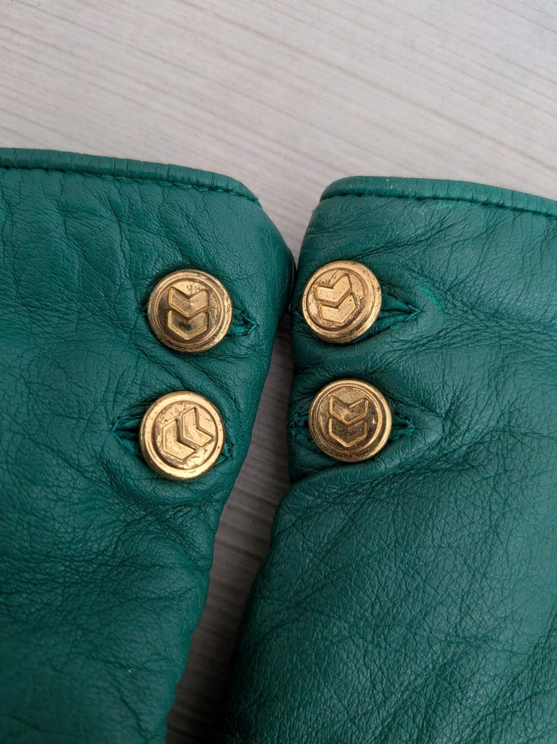 May include: Close-up of a pair of emerald green leather gloves. Each glove has three gold-colored buttons with a stylized arrow design. The gloves are made of a soft, textured leather.