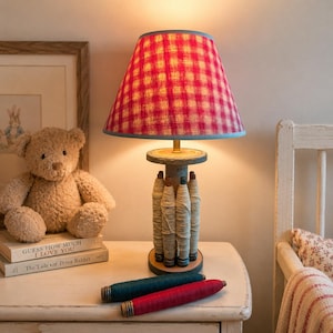 May include: A table lamp with a red and white checkered fabric lampshade and a base constructed from a wooden spool wrapped with beige yarn. Two additional yarn bobbins in dark teal and red lie horizontally on the surface in front of the lamp.