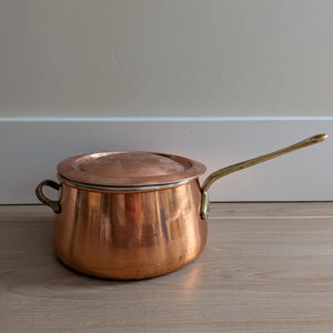 May include: A round copper saucepan with a matching lid, featuring a long brass handle and a small brass side handle. The exterior surface of the pot shows a polished metallic finish with subtle signs of use and natural patina.