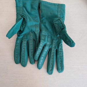 May include: A pair of emerald green gloves. The gloves are made of a soft material, with perforated details on the fingers and a stitched seam along the back. The gloves have a classic design.