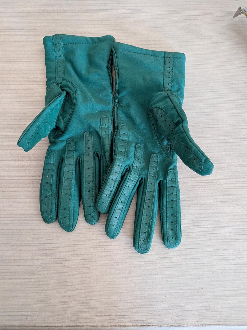 May include: A pair of emerald green gloves. The gloves are made of a soft material, with perforated details on the fingers and a stitched seam along the back. The gloves have a classic design.