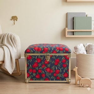 May include: A rectangular storage trunk upholstered in a dark blue fabric featuring a repeating pattern of red apples and green leaves. The trunk has metallic gold-toned corner reinforcements and a central latch closure on the front panel.