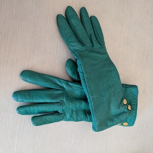 May include: A pair of emerald green leather gloves with gold-colored button details on the cuffs. The gloves are laid out on a light-colored wooden surface, showcasing their smooth texture and classic design.