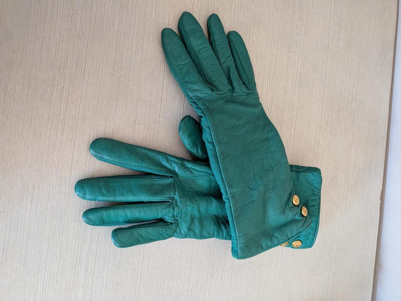 May include: A pair of emerald green leather gloves with gold-colored button details on the cuffs. The gloves are laid out on a light-colored wooden surface, showcasing their smooth texture and classic design.