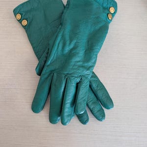 May include: A pair of emerald green leather gloves. Each glove has two gold-colored buttons on the cuff. The gloves are made of soft, supple leather and appear to be unworn.