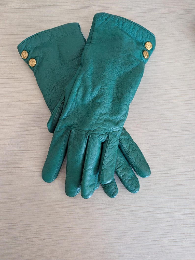 May include: A pair of emerald green leather gloves. Each glove has two gold-colored buttons on the cuff. The gloves are made of soft, supple leather and appear to be unworn.