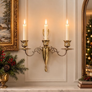 May include: A polished brass wall sconce featuring three arms with candle cups, each holding a lit white taper candle. The fixture has a central vertical mount with decorative scrollwork extending to the two side candle holders.
