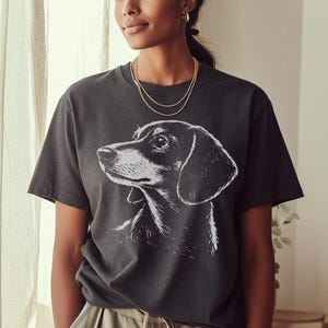 May include: Dark gray t-shirt featuring a black and white illustration of a dachshund dog. The dog's profile is visible, with detailed shading. The shirt has a classic crew neck and short sleeves, suitable for casual wear.