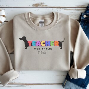 Pode incluir: Camisola bege com a palavra "TEACHER" em letras coloridas, integrada com uma silhueta de dachshund preto. O texto "MRS. ADAMS 3rd Grade" est&aacute; abaixo. A camisola &eacute; combinada com jeans azuis.