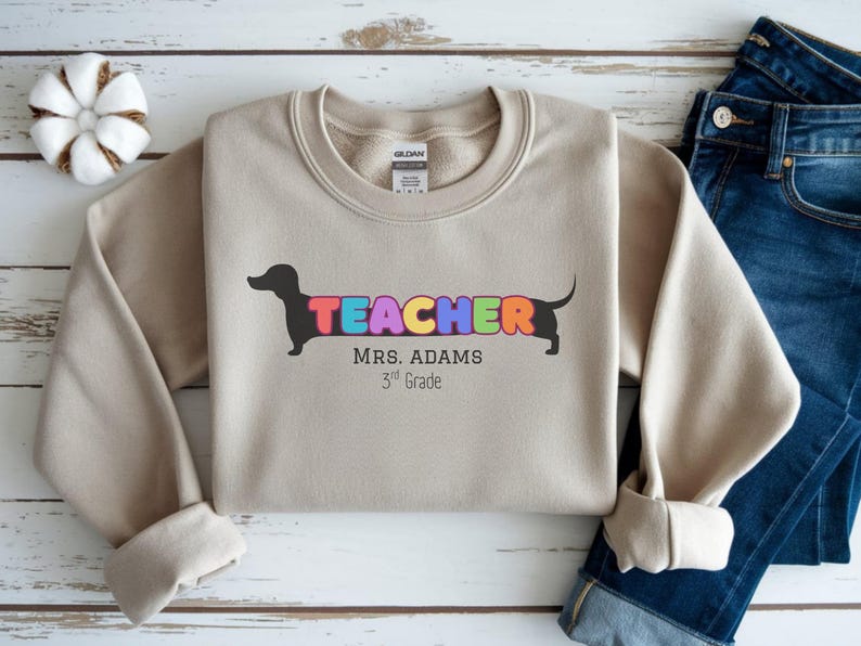 Pode incluir: Camisola bege com a palavra "TEACHER" em letras coloridas, integrada com uma silhueta de dachshund preto. O texto "MRS. ADAMS 3rd Grade" est&aacute; abaixo. A camisola &eacute; combinada com jeans azuis.
