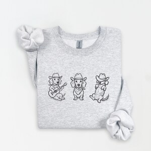 May include: A light gray sweatshirt featuring three dachshund illustrations in cowboy hats. One dog plays a guitar, another stands, and the third sits. The illustrations are in black ink.