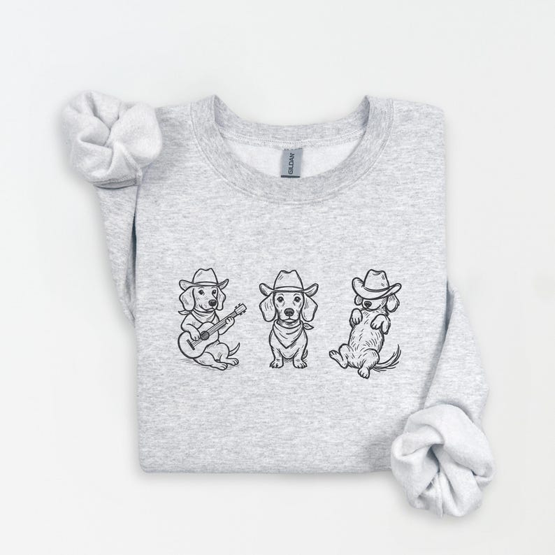 May include: A light gray sweatshirt featuring three dachshund illustrations in cowboy hats. One dog plays a guitar, another stands, and the third sits. The illustrations are in black ink.