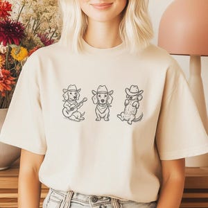 May include: Beige t-shirt featuring a graphic of three dachshunds wearing cowboy hats. One dog is playing a guitar, another is wearing a bandana, and the third is lounging. The design is in black ink.