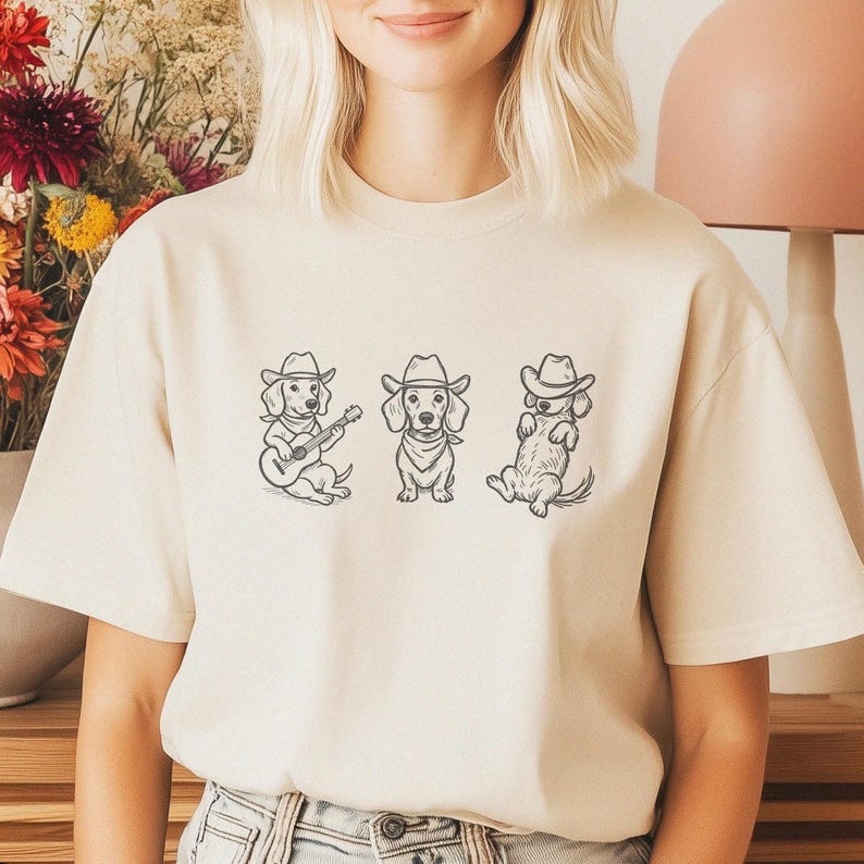 May include: Beige t-shirt featuring a graphic of three dachshunds wearing cowboy hats. One dog is playing a guitar, another is wearing a bandana, and the third is lounging. The design is in black ink.