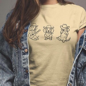 May include: Tan t-shirt featuring three dachshund illustrations in cowboy hats. One dog plays a guitar, another sits upright, and the third reclines. The design is in black ink. The model is wearing a denim jacket.