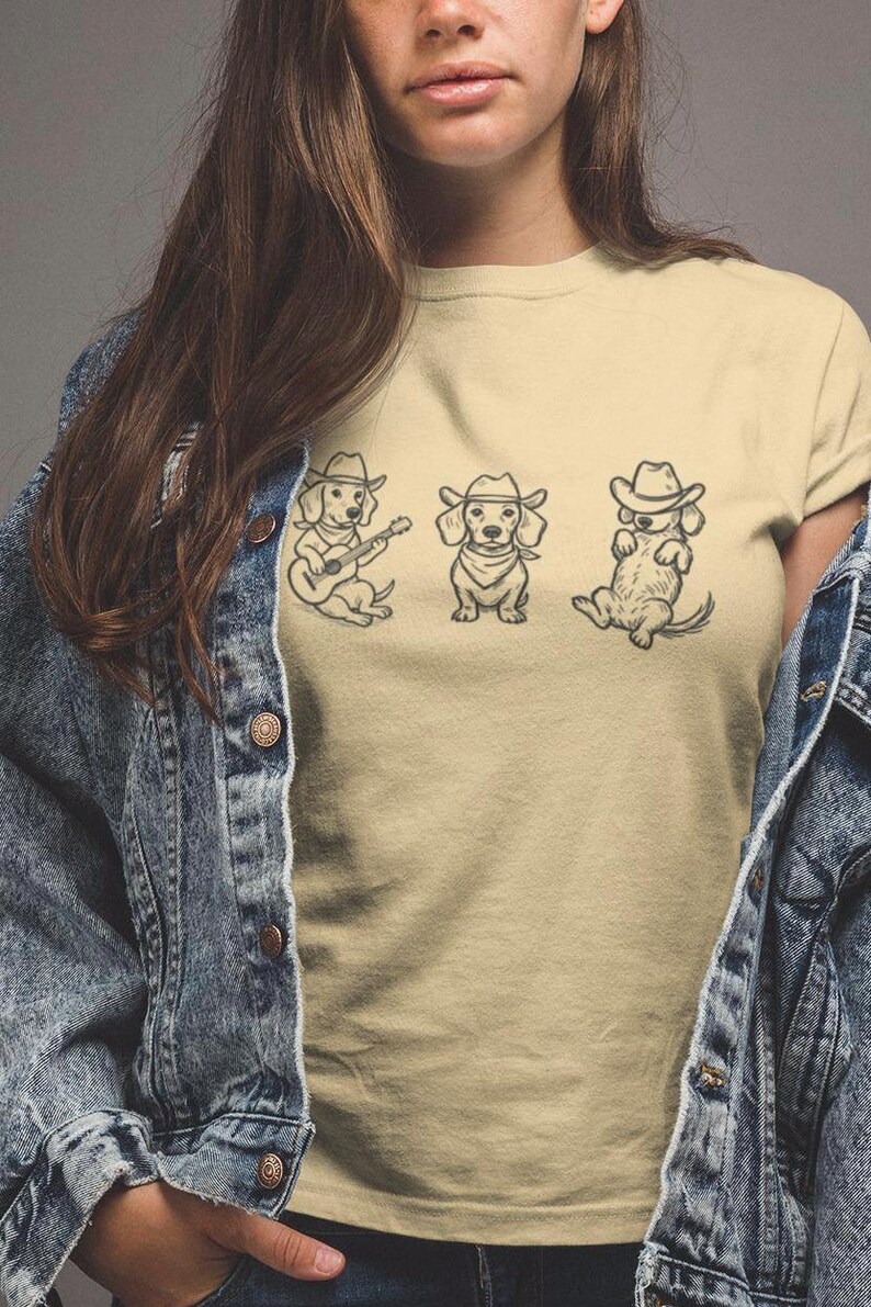 May include: Tan t-shirt featuring three dachshund illustrations in cowboy hats. One dog plays a guitar, another sits upright, and the third reclines. The design is in black ink. The model is wearing a denim jacket.