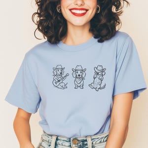 May include: A light blue t-shirt featuring three dachshunds wearing cowboy hats. One dog plays a guitar, another wears a bandana, and the third sits upright. The graphic is in black ink.