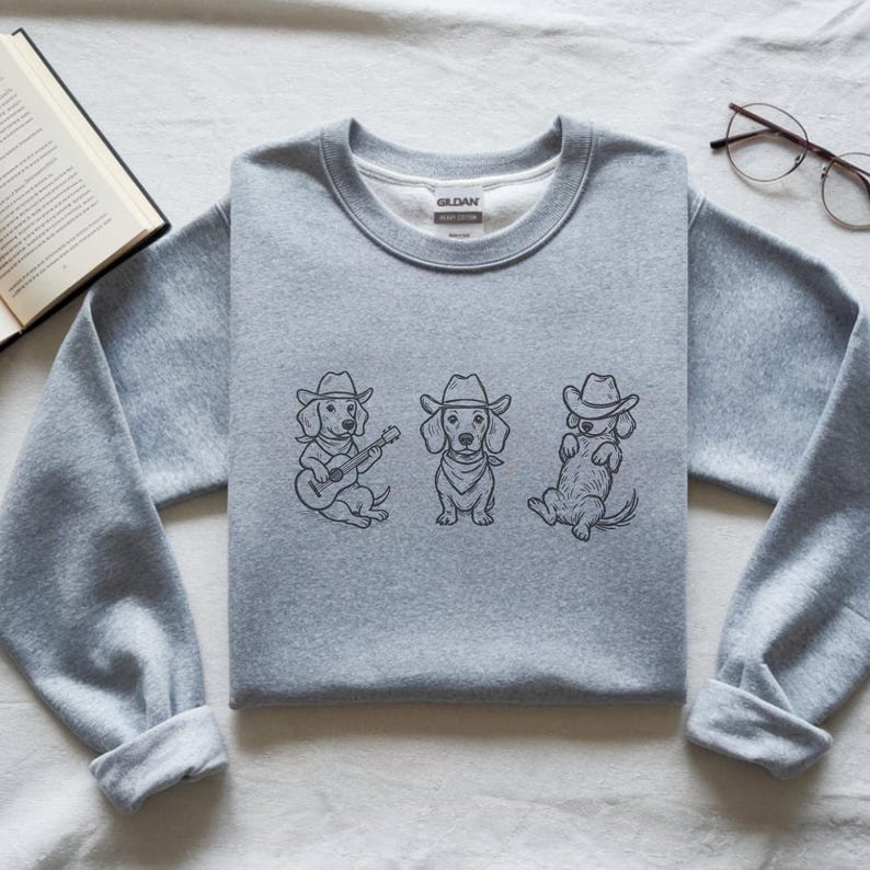 May include: A light gray sweatshirt featuring a line drawing of three dachshunds wearing cowboy hats. One dog plays a guitar, another wears a bandana, and the third reclines. The sweatshirt has a crew neck and long sleeves.