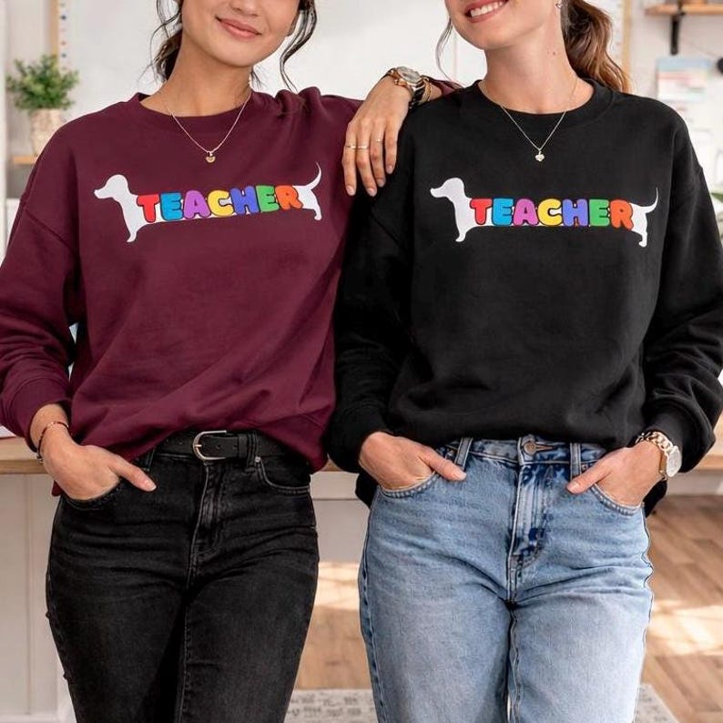 Pode incluir: Duas camisolas, uma bord&ocirc; e outra preta, cada uma com a silhueta de um c&atilde;o dachshund branco e a palavra "TEACHER" em cores do arco-&iacute;ris. As camisolas s&atilde;o de gola redonda.
