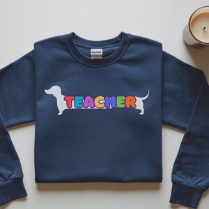 Pode incluir: Camisola azul marinho com um desenho de c&atilde;o dachshund branco e a palavra "TEACHER" em letras coloridas. Uma vela acesa num frasco de vidro est&aacute; no canto superior direito. A camisola tem mangas compridas e gola redonda.