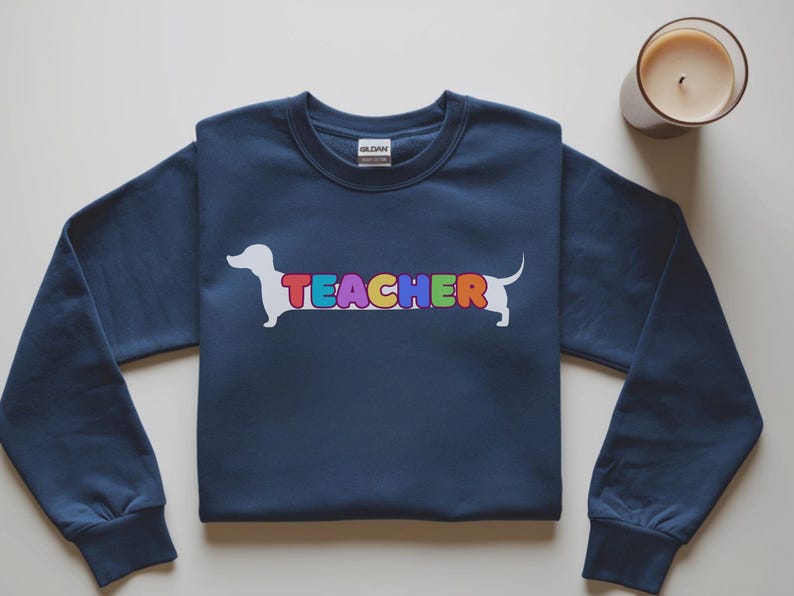Pode incluir: Camisola azul marinho com um desenho de c&atilde;o dachshund branco e a palavra "TEACHER" em letras coloridas. Uma vela acesa num frasco de vidro est&aacute; no canto superior direito. A camisola tem mangas compridas e gola redonda.