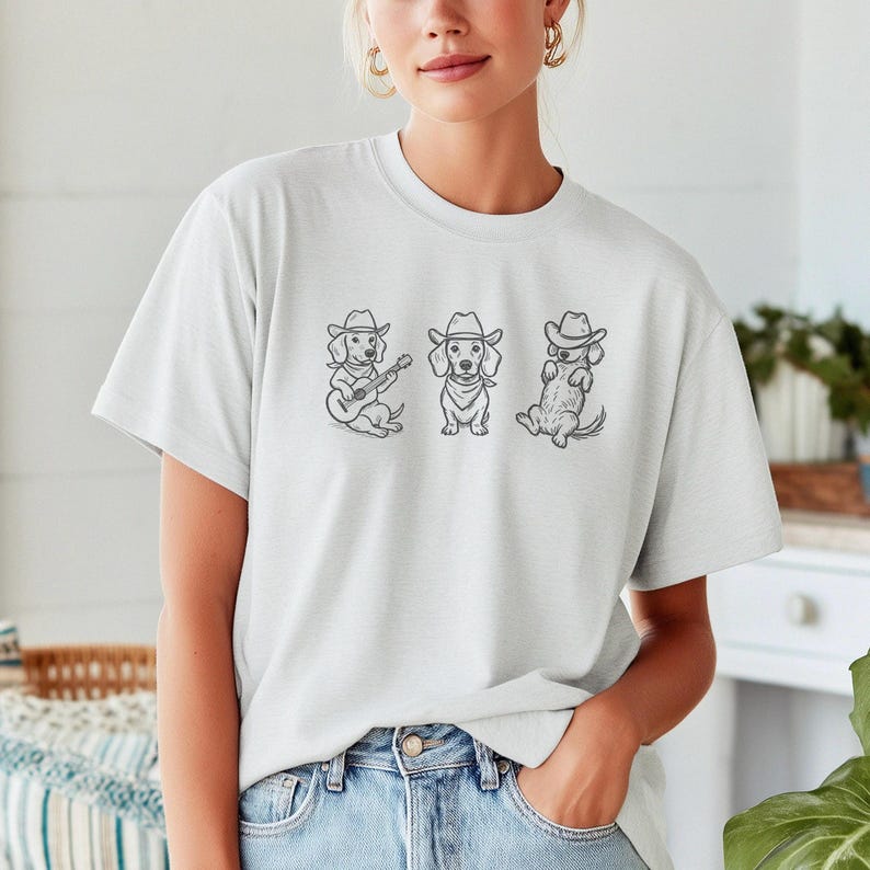May include: A light gray t-shirt featuring three dachshunds wearing cowboy hats. One dog plays a guitar, another wears a bandana, and the third sits up. The design is in black ink.