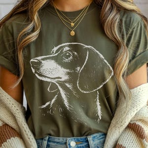May include: Olive green t-shirt featuring a detailed white illustration of a dachshund's head and shoulders. The dog's profile is facing left, with a focus on its eye and ear. The shirt has rolled-up sleeves and is paired with blue jeans.