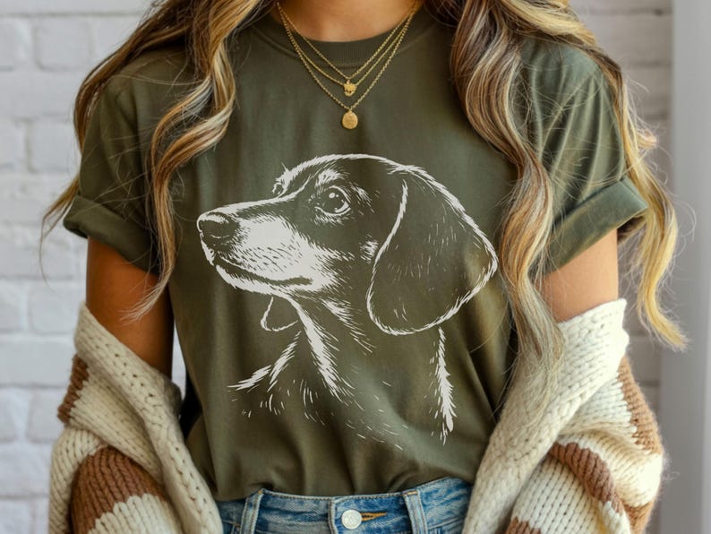 May include: Olive green t-shirt featuring a detailed white illustration of a dachshund's head and shoulders. The dog's profile is facing left, with a focus on its eye and ear. The shirt has rolled-up sleeves and is paired with blue jeans.