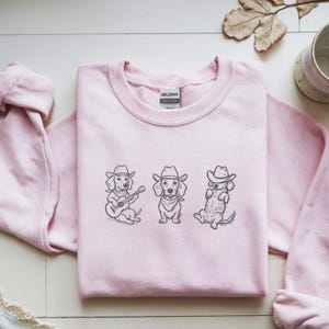 May include: A light pink sweatshirt featuring three dachshunds wearing cowboy hats. The dachshunds are illustrated in black and white, with one playing a guitar, one standing, and one lying down. The sweatshirt has a crew neck.