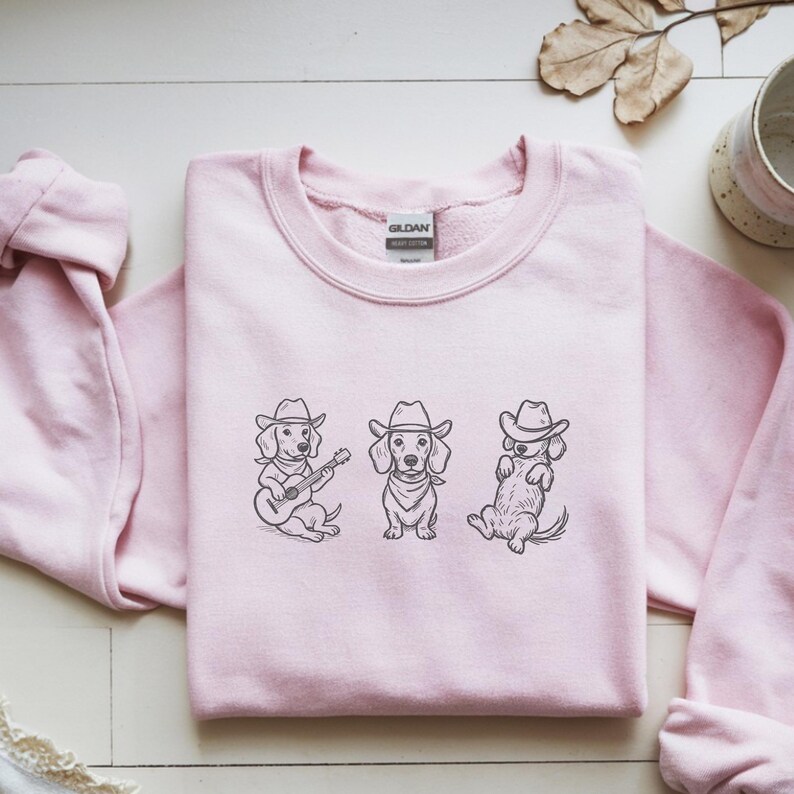 May include: A light pink sweatshirt featuring three dachshunds wearing cowboy hats. The dachshunds are illustrated in black and white, with one playing a guitar, one standing, and one lying down. The sweatshirt has a crew neck.