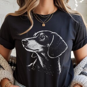 May include: Black t-shirt featuring a detailed, black and white illustration of a dachshund dog's head in profile. The dog's features are clearly defined, with a focus on its alert expression. The shirt is a classic crew neck style.