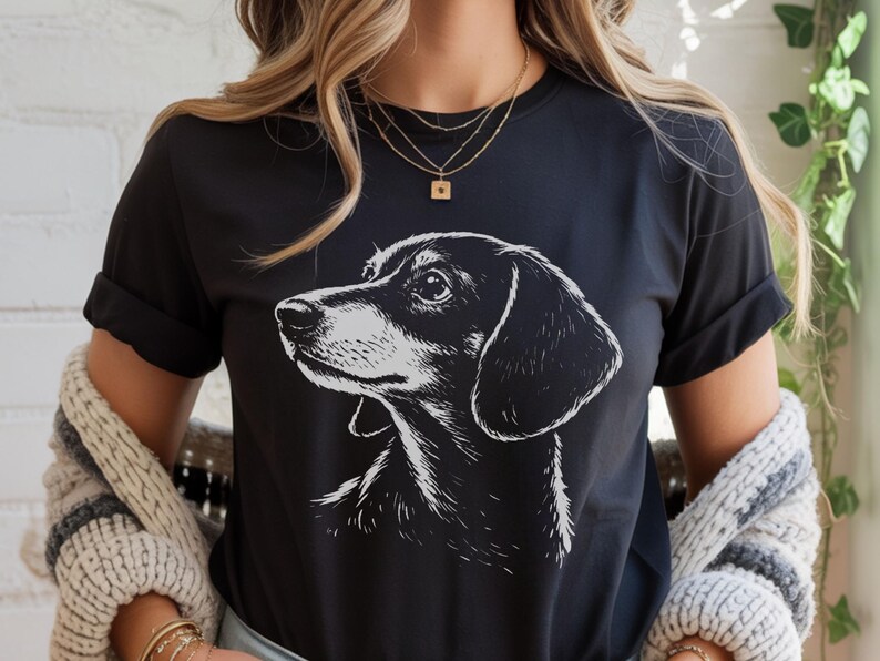 May include: Black t-shirt featuring a detailed, black and white illustration of a dachshund dog's head in profile. The dog's features are clearly defined, with a focus on its alert expression. The shirt is a classic crew neck style.