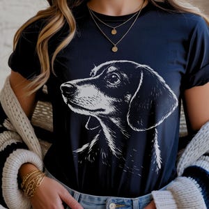 May include: Navy blue t-shirt featuring a black and white illustration of a dachshund dog's head. The dog is facing left, with detailed shading. The shirt has short sleeves and a round neckline.