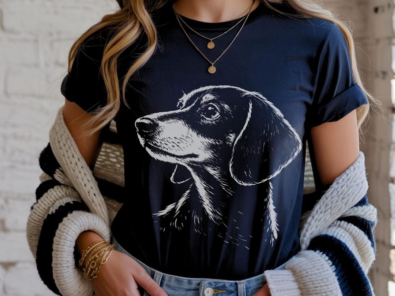 May include: Navy blue t-shirt featuring a black and white illustration of a dachshund dog's head. The dog is facing left, with detailed shading. The shirt has short sleeves and a round neckline.