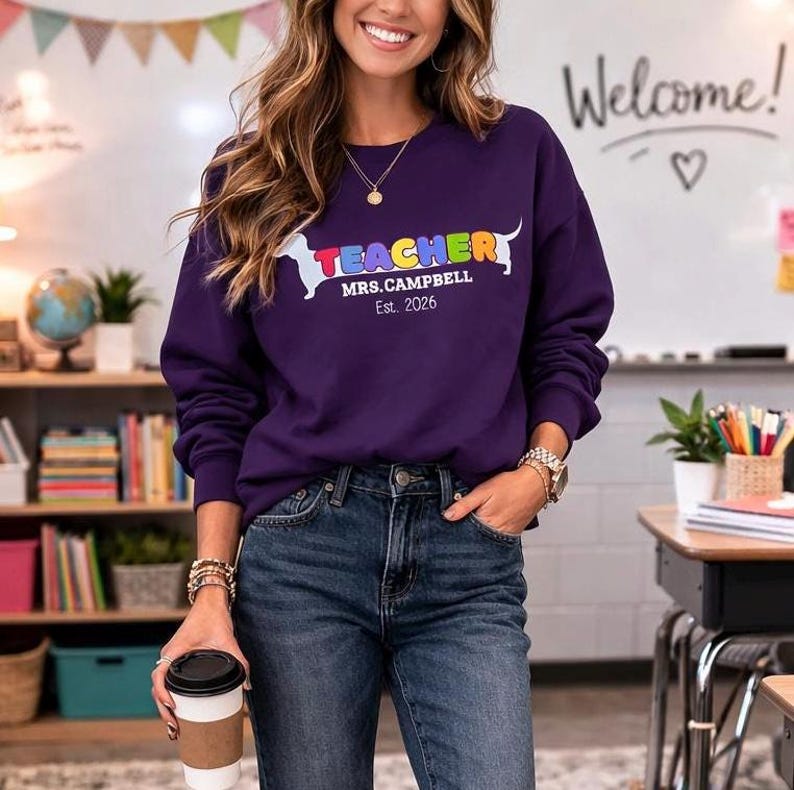 Pode incluir: Um moletom roxo com a palavra "TEACHER" em letras coloridas e um gr&aacute;fico de cachorro dachshund. Abaixo da palavra "TEACHER" est&aacute; o texto "MRS.CAMPBELL Est. 2026". A pessoa est&aacute; usando jeans azuis e segurando uma x&iacute;cara de caf&eacute;.