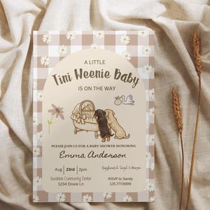 May include: A rectangular baby shower invitation card featuring a beige and white gingham background with white daisy patterns. The centre has an arched cream panel with the text A LITTLE Tini Weenie Baby IS ON THE WAY, featuring illustrations of two dachshunds looking into a bassinet, a stork, and a flower. The card includes event details for Emma Anderson on August 23rd at 2pm at the Sunshine Community Center.