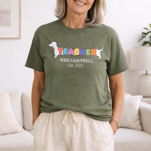 Pode incluir: T-shirt verde-oliva com um gr&aacute;fico de c&atilde;o dachshund e a palavra "TEACHER" em cores do arco-&iacute;ris. Abaixo do gr&aacute;fico est&aacute; escrito "MRS. CAMPBELL Est. 2026". A camiseta &eacute; usada por uma pessoa.