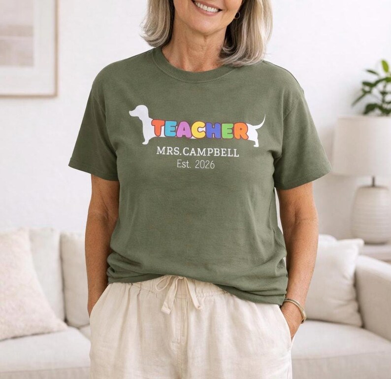 Pode incluir: T-shirt verde-oliva com um gr&aacute;fico de c&atilde;o dachshund e a palavra "TEACHER" em cores do arco-&iacute;ris. Abaixo do gr&aacute;fico est&aacute; escrito "MRS. CAMPBELL Est. 2026". A camiseta &eacute; usada por uma pessoa.