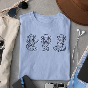 May include: Light blue t-shirt featuring three dachshunds in cowboy hats. One dog plays a guitar, another stands, and the third reclines. The shirt is surrounded by accessories including a camera, sunglasses, and a hat.