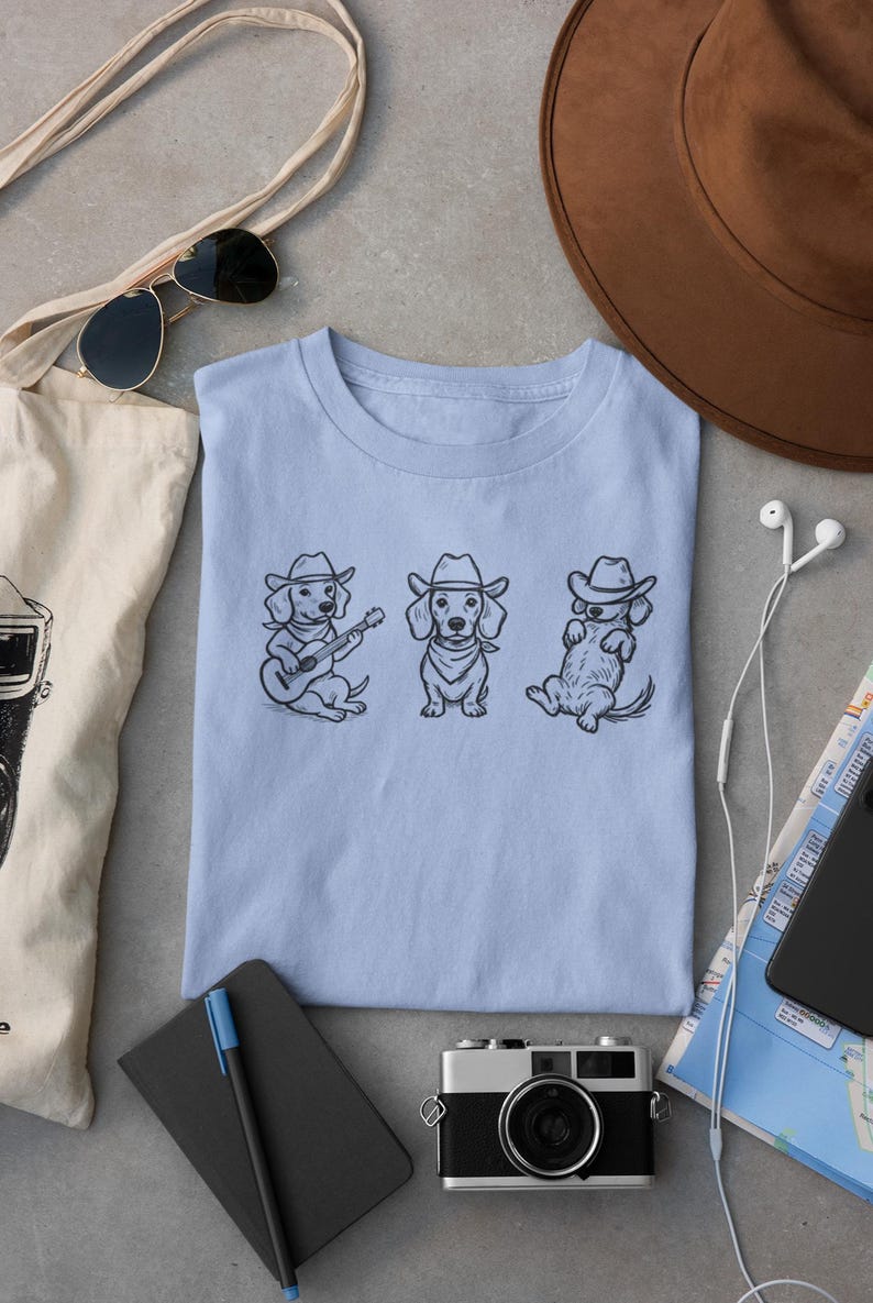 May include: Light blue t-shirt featuring three dachshunds in cowboy hats. One dog plays a guitar, another stands, and the third reclines. The shirt is surrounded by accessories including a camera, sunglasses, and a hat.