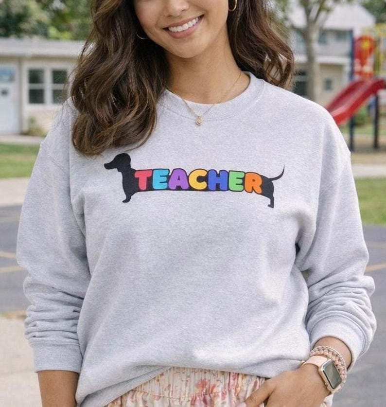 Pode incluir: Camisola cinza claro com a palavra "TEACHER" em letras coloridas do arco-&iacute;ris, integrada na silhueta de um c&atilde;o dachshund. A camisola &eacute; de estilo casual, adequada para uso di&aacute;rio.