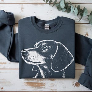May include: A dark gray sweatshirt featuring a white illustration of a dachshund's head. The dog's profile is detailed, with a focus on its eye and ear. The sweatshirt is laid flat on a light wood surface.