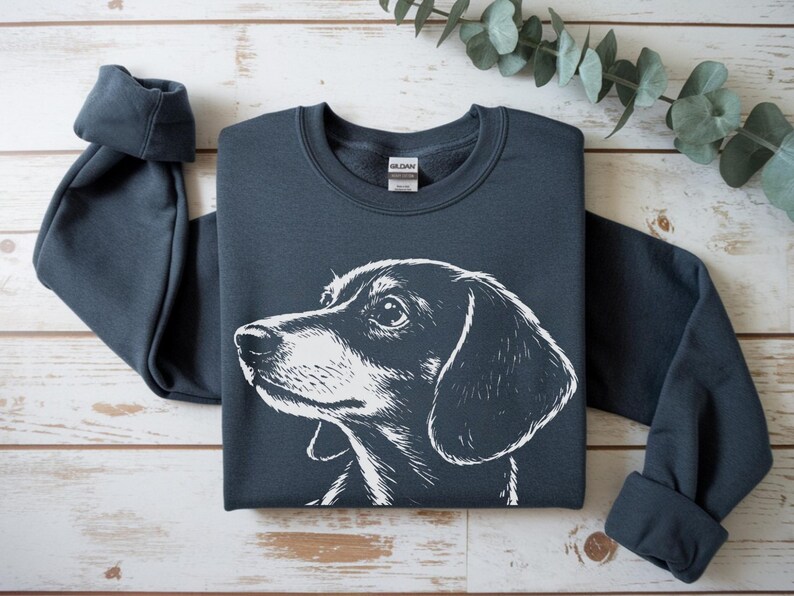 May include: A dark gray sweatshirt featuring a white illustration of a dachshund's head. The dog's profile is detailed, with a focus on its eye and ear. The sweatshirt is laid flat on a light wood surface.