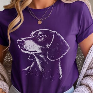 May include: Purple t-shirt featuring a detailed white illustration of a Dachshund dog's head and shoulders. The dog's features are finely rendered, with a focus on the eyes and ears. The shirt has rolled-up sleeves.