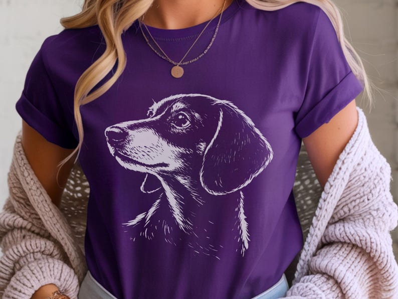 May include: Purple t-shirt featuring a detailed white illustration of a Dachshund dog's head and shoulders. The dog's features are finely rendered, with a focus on the eyes and ears. The shirt has rolled-up sleeves.