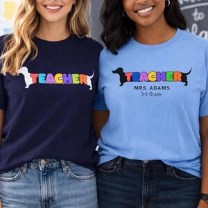 Pode incluir: Duas mulheres vestindo camisetas combinando. Uma camisa &eacute; azul marinho, a outra azul claro. Ambas as camisas t&ecirc;m a palavra "TEACHER" em letras coloridas com um gr&aacute;fico de cachorro dachshund. A camisa azul marinho tem um cachorro branco, a camisa azul claro tem um cachorro preto.