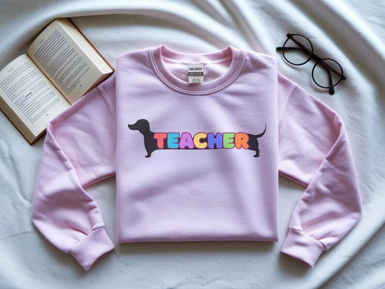 Pode incluir: Camisola rosa claro com a palavra "TEACHER" em letras coloridas e um desenho de c&atilde;o dachshund preto. Um par de &oacute;culos pretos e um livro aberto tamb&eacute;m est&atilde;o na imagem.
