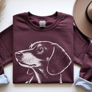 May include: A maroon sweatshirt featuring a white illustration of a dachshund dog's head. The sweatshirt has a crew neck and cuffed sleeves with a light blue lining. A brown hat and a cup of coffee are in the background.