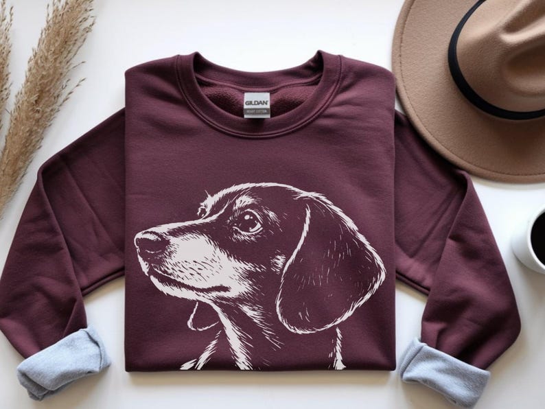 May include: A maroon sweatshirt featuring a white illustration of a dachshund dog's head. The sweatshirt has a crew neck and cuffed sleeves with a light blue lining. A brown hat and a cup of coffee are in the background.