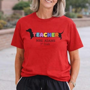 Pode incluir: Camiseta vermelha com a palavra &laquo;TEACHER&raquo; em letras coloridas, desenhada como um c&atilde;o dachshund. Abaixo, o texto &laquo;MRS. ADAMS 3rd Grade&raquo; em preto. A camiseta &eacute; de gola redonda.